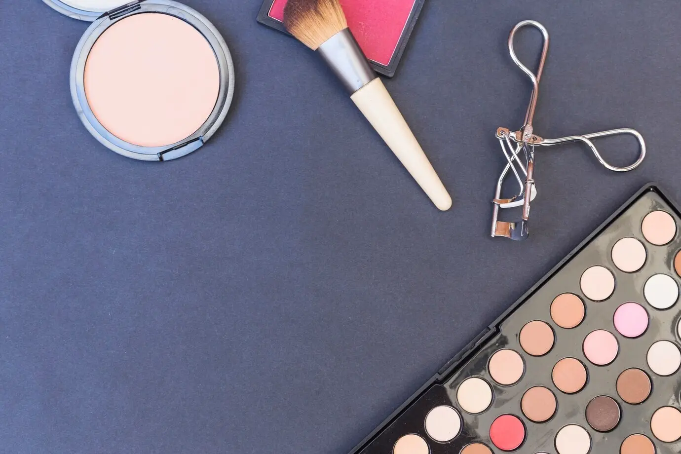 A top-down view of a makeup brush, blusher, eyeshadow palette, and eyelash curler on a blue backdrop.